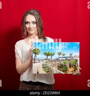 Fotoleinwand. Eine lächelnde Frau hält eine rechteckige Fotografie mit Galerie Wrap auf rotem Hintergrund. Landschaftsfoto auf glänzendem Kunststoff gedruckt Stockfoto