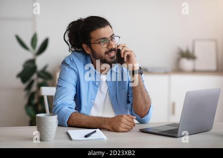 Millennial Indian Man Im Gespräch Auf Handy Und Mit Laptop An Heimbüro Stockfoto