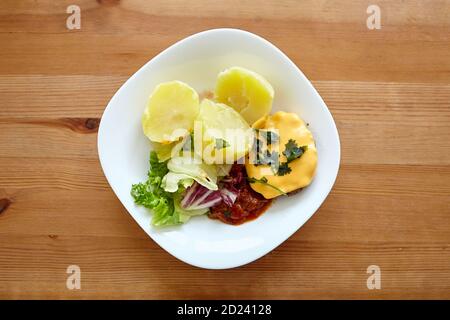 Gekochte Kartoffeln mit grünem Salat, Gemüse und Kotelet. Weißer Teller mit hausgemachtem Essen auf einem hölzernen Esstisch. Gekochte Kartoffelschale auf Holzbackgro Stockfoto