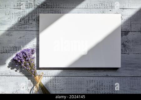 Weiße leere Leinwand und Strauß von Lavendelblüten auf grauem Holztisch mit einem Schatten von Sonnenlicht bedeckt. Modell, Draufsicht Stockfoto