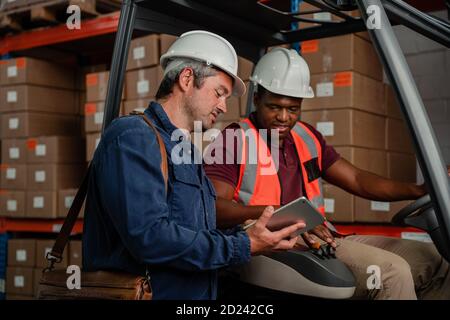 Zwei männliche Arbeiter tragen Harthüte halten digitale Tablette Recherche Standort des Paketes, während der Fahrt Fabrikkarte im Lager. Stockfoto