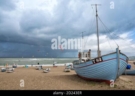 Sturmwolken über dem Nordseestrand von Nørre Vorupør, Dänemark, mit Fischerbooten auf dem Sand und Kitesurfern in den Wellen Stockfoto