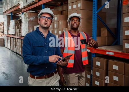 Ein Geschäftsmann trägt eine Brille mit einem digitalen Tablet, während ein Fabrikarbeiter den Vorgesetzten im Lager zeigt. Stockfoto