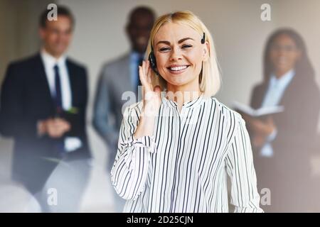 Junge Geschäftsfrau mit Headset und lächelnd im Stehen Ein Büro mit diversen Mitarbeitern im Hintergrund Stockfoto