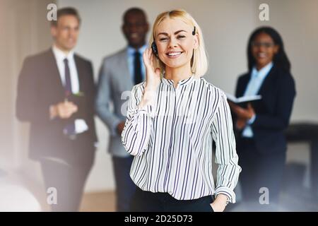 Junge Geschäftsfrau mit Headset und lächelnd im Stehen Ein Büro mit diversen Kollegen, die im Hintergrund arbeiten Stockfoto