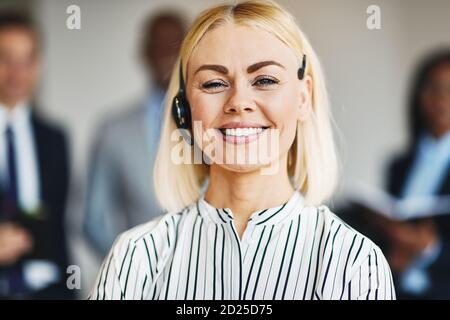 Lächelnde junge Geschäftsfrau, die ein Headset im Büro trägt Mit einer vielfältigen Gruppe von Kollegen im Hintergrund Stockfoto