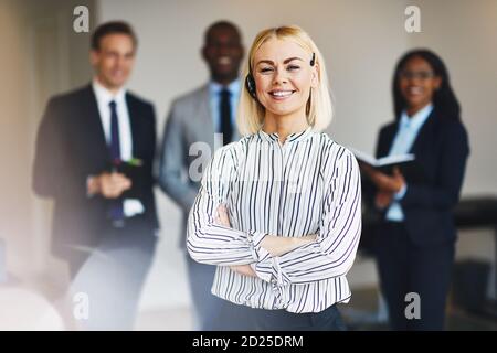 Lächelnde junge Geschäftsfrau, die ein Headset im Büro trägt Mit einer Gruppe verschiedener Kollegen im Hintergrund Stockfoto