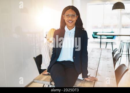 Lächelnde junge afrikanische Geschäftsfrau sitzt auf einem Sitzungstisch mit Eine Gruppe von Kollegen, die im Hintergrund arbeiten Stockfoto