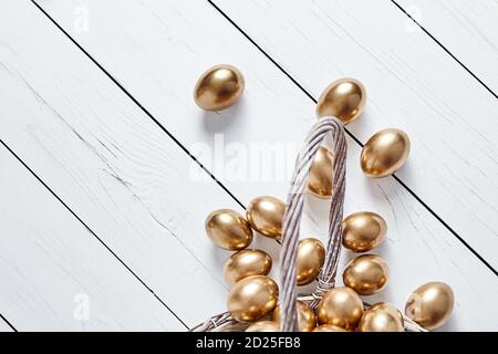 Goldene Ostereier verschüttet aus Korbkorb auf weißem Holztisch Hintergrund. Draufsicht Stockfoto