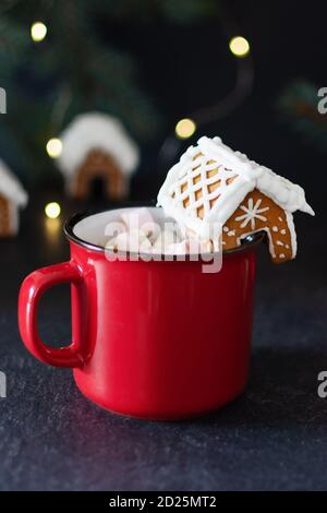 Roter Becher heißer Schokolade mit Marshmallow und hausgemachtem Lebkuchen kleines Haus zur Dekoration auf schwarzem Hintergrund im weihnachtlichen Interieur. Weihnachtsurlaub Stockfoto