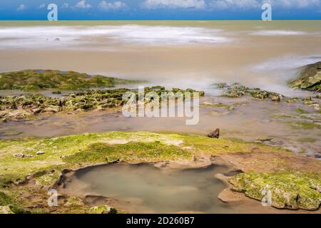 Verschwommenes Meer über Riffen mit Algen und unter blauem Himmel mit Nimbus-Wolken aufgeladen. Stockfoto