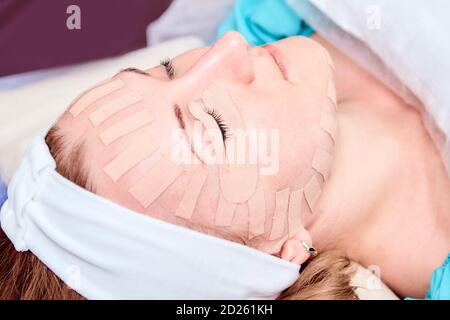 Kinesiotaping. Junge Frau liegt mit Klebeband auf ihrem Gesicht. Physiotherapie und Kosmetologie Verfahren. Methode der nicht-chirurgischen Hautverjüngung. Gesichtshaut Stockfoto