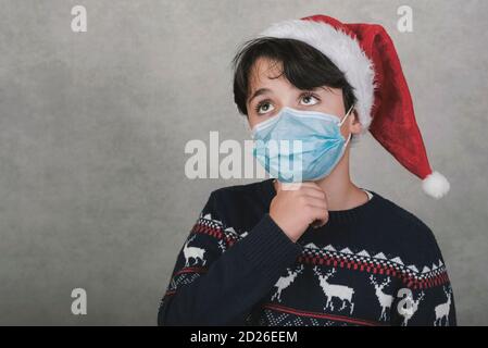 Frohe Weihnachten, nachdenkliches Kind mit medizinischer Maske trägt Weihnachtsmann Hut Auf grauem Hintergrund Stockfoto