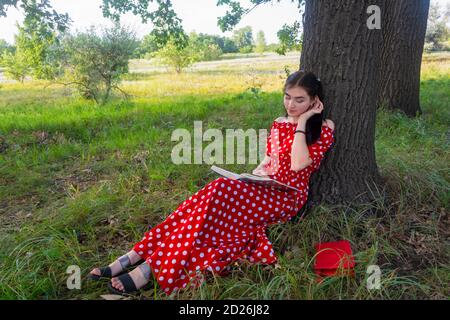 Eine schöne Brünette in einem langen roten Kleid im Sommer liest ein Buch in einem Park. Ein Schüler bereitet sich auf den Unterricht vor. Sommerstimmung, Harmonie, Erholung im Freien. Stockfoto