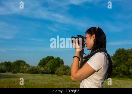 Eine schöne Brünette Fotografin schaut in ihre SLR-Kamera und fotografiert die Natur mit einer Digitalkamera. Sommer strahlend blauer Himmel und Wiese mit grünen Bäumen Stockfoto