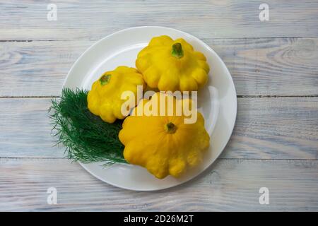 Gelbe Mini-Patty Pfanne Squash auf einem weißen Teller neben grünen Dill. Zucchini auf Holzhintergrund, selektiver Fokus, Nahaufnahme. Stockfoto