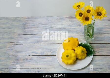 Stillleben auf einem Holztisch im Herbst. Gelbe Blüten in einer Glasvase und runde gelbe Zucchini auf einem Teller. Nahaufnahme, selektiver Fokus. Stockfoto