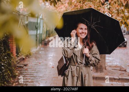 Junge hübsche Frau Porträt Musik hören mit Headset beim Gehen Auf der Straße mit Regenschirm bei Regen Stockfoto