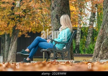 Blonde Mädchen in einer türkisfarbenen Jacke und blaue Jeans sitzt auf einer Bank in einem Herbst-Park, auf dem Boden gefallen gelben Blättern Stockfoto