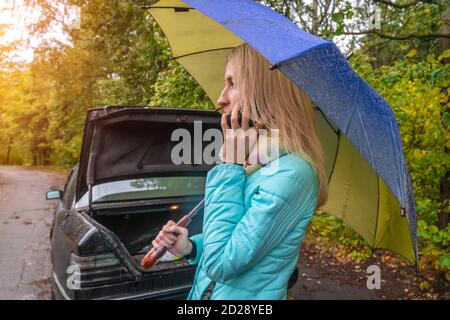 Ein schlankes blondes Mädchen steht in der Nähe eines kaputten Autos mit Ein Regenschirm in ihren Händen und spricht am Telefon Und bittet um Hilfe Stockfoto