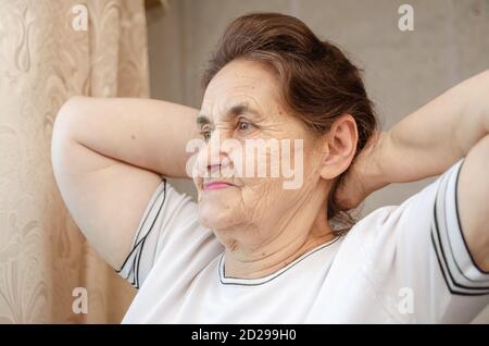 Ältere dunkelhaarige Frau mit ihren Händen hinter ihrem Kopf Schaut aus dem Fenster Stockfoto