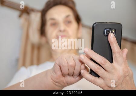 Ältere Frau mit dunklem Haar hält ein schwarzes Handy vor sich, schaut es an und tippt mit dem Finger auf den Bildschirm Stockfoto