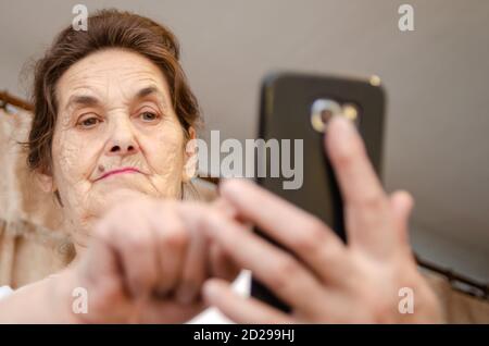 Ältere Frau mit dunklem Haar hält ein schwarzes Handy vor sich, schaut es an und tippt mit dem Finger auf den Bildschirm Stockfoto