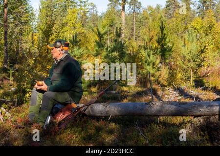 Männlicher Elchjäger, der sich auf einem umgestürzten Baum mit einem alten handgemachten Holzbecher in der Hand ausruhte, Foto aus Brattforsmen Vasternorrland Schweden. Stockfoto