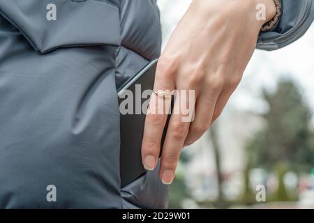 Die Hand der Frau mit einem Ring zieht ein schwarzes Smartphone heraus Von der Tasche einer grauen Jacke Nahaufnahme Stockfoto