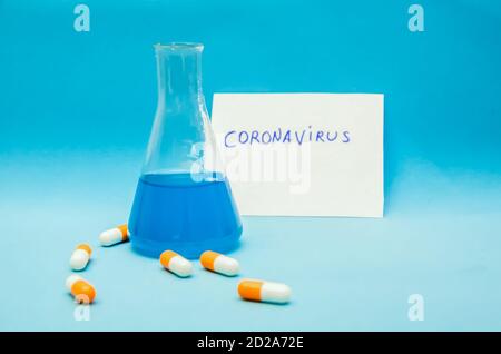 Flasche mit blauer Flüssigkeit, orange-weiße Kapseln auf blauem Hintergrund, im Hintergrund ein weißes Blatt Papier mit der Aufschrift Coronavirus Stockfoto