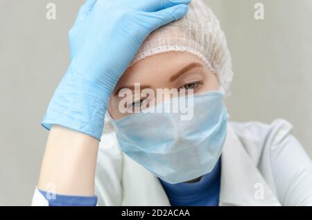 Porträt einer schönen jungen Ärztin in einem medizinischen Kleid, medizinische Maske und Mütze mit dem Kopf auf der Hand gestützt. Müder Arzt Stockfoto