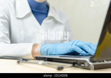 Arzt in einem medizinischen Kleid, blaue medizinische Handschuhe arbeiten an einem Laptop, neben einem Stethoskop. Konzept der Medizin Stockfoto