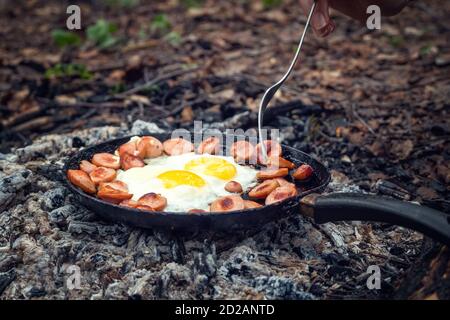 Mädchenhand mit einer Gabel dreht sich über Stücke von gebratener Wurst und Eier in einer Pfanne auf die Kohlen. Frühstück in der Natur, Picknick, Campingkost. Stockfoto