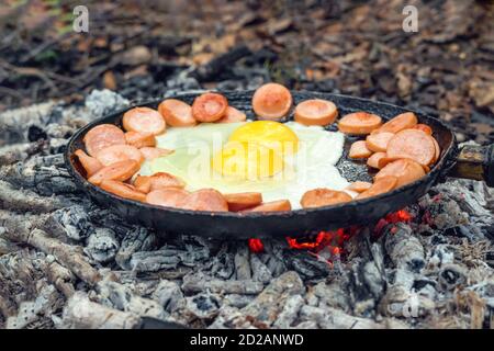 Zwei Eier und Würstchen werden in einer Pfanne auf Kohlen im Wald gebraten. Frühstück in der Natur, Picknick. Stockfoto