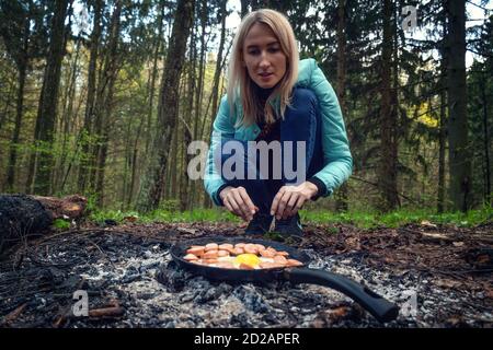 Junge blonde Frau röstet Eier und Würstchen auf Kohlen im Wald bei gutem Sommerwetter. Konzept von Kampagnen. Frühstück in der Natur. Stockfoto