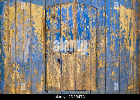 Alte Holztür mit blätternder blauer Farbe überzogen. Eine gelbe Holzwand und eine halbrunde arabische Tür mit Klopfgriff. Stockfoto