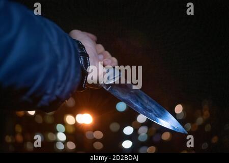 Nahaufnahme der Hand eines Mannes mit einem Messer in der Nacht auf der Straße zwischen den Lichtern der Häuser. Konzept der Kriminalität, Raub Stockfoto