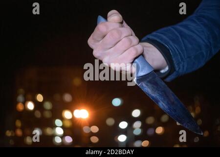 Nahaufnahme der Hand eines Mannes mit einem Messer in der Nacht auf der Straße zwischen den Lichtern der Häuser. Konzept der Kriminalität, Raub Stockfoto