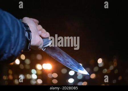 Nahaufnahme der Hand eines Mannes mit einem Messer in der Nacht auf der Straße zwischen den Lichtern der Häuser. Konzept der Kriminalität, Raub Stockfoto