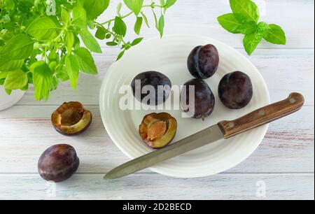 Reife frische Pflaumen und Messer mit Holzgriff Ein weißer Teller und Küchenkräuter auf lackierter Holzoberfläche Stockfoto