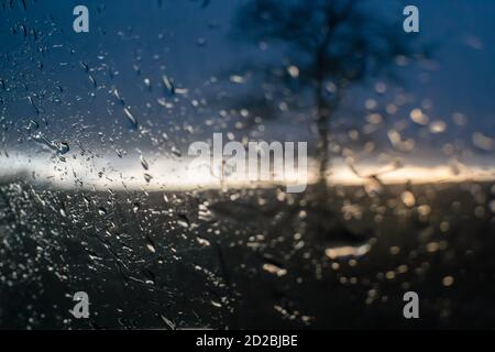 Regentropfen auf dem Seitenfenster des Autos vor dem Hintergrund des Sonnenuntergangs und der Silhouette eines Baumes, ein Foto aus dem Inneren des Autos Stockfoto