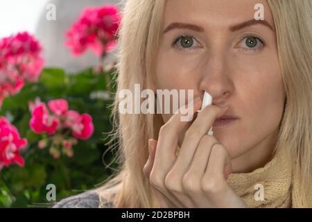 Junge blonde Frau verwendet ein Nasenspray für eine laufende Nase oder Allergie vor einem Hintergrund von Innenblumen, kopieren Raum. Stockfoto