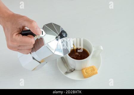 Eine Frauenhand hält eine Geysir-Kaffeemaschine. Schwarzer heißer Kaffee gießt in eine weiße Porzellanschale. Stockfoto