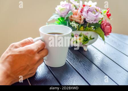 Eine Männerhand hält eine weiße Tasse Kaffee auf einem Holztisch in der Nähe, neben einem Blumenstrauß Stockfoto