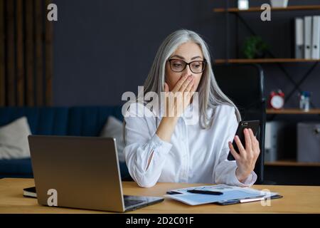 Überrascht Senior grauhaarige schöne Frau hält Smartphone. Glückliche Geschäftsfrau bekam schockierende positive Nachrichten. Verwenden von Handy-Apps, Einkaufen Stockfoto