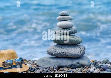 Ein Steinhaufen in der Nähe des Meeres, neben einem Strohhut und Gläsern. Steinturm am Strand an einem sonnigen Tag. Gleichgewicht, Seelenfrieden. Stockfoto