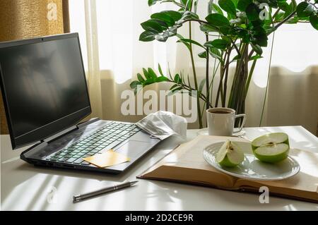Ein Arbeitsplatz mit einem Computer, einem offenen Buch, einem Apfel, einer medizinischen Maske und einer Tasse Kaffee.
