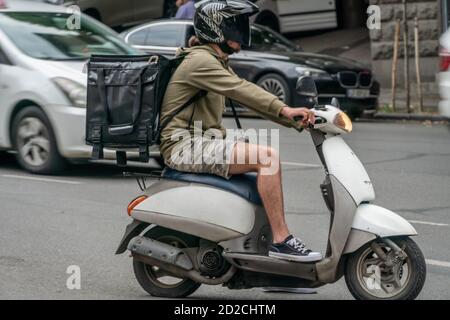 Kurier mit einem Rucksack auf dem Rücken auf einem weißen Roller mit Lebensmittelzustellung auf der Straße Stockfoto