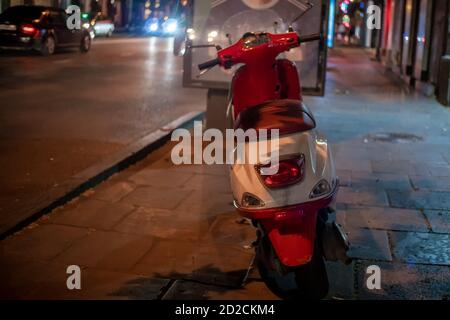 Moderne Roller auf dem Bürgersteig in der Nähe der Straße am Abend. Ein rot-weißes Moped steht nachts am Straßenrand. Stockfoto