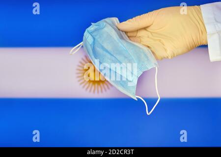 Medizinische Maske in den Händen eines Arztes und die Argentinien-Flagge, Kopierraum. Schutzhandschuh mit Maske Nahaufnahme auf dem Hintergrund des Staates Argentinien Stockfoto
